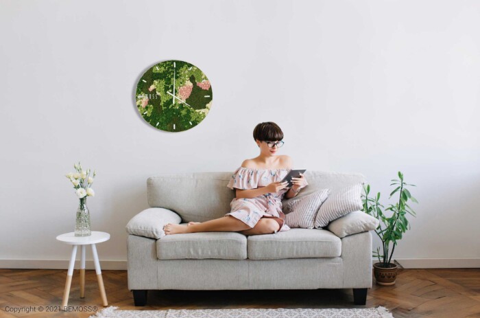 Eine Frau sitzt auf einer Couch mit einem Buch in der Hand und einer Uhr im Hintergrund mit einem Vogel darauf, moosbild, mooswand, moos pflanzen, moos, moos deko, moos art